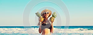 Summer vacation, beautiful happy smiling woman in black bikini swimsuit and straw hat holding pineapple fruits on the beach on sea