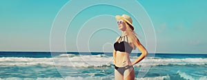 Summer vacation, beautiful happy smiling woman looking away in bikini swimsuit and straw hat on the beach on sea coast with waves