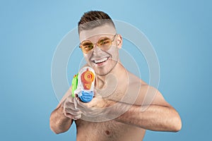 Summer vacation concept. Attractive millennial man pointing water gun at screen over blue studio background