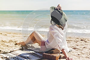 Summer vacation concept. Happy young woman relaxing on beach. Hipster slim girl in white shirt and hat sitting and tanning on