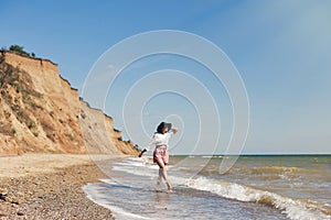 Summer vacation concept. Happy young woman in white shirt and hat walking on sunny beach. Hipster slim girl relaxing near sea with