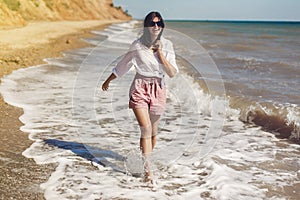 Summer vacation concept. Happy young woman in white shirt and sunglasses running and having fun with waves on sunny beach. Hipster
