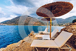 Summer vacation destination. Straw sunshades and sunbeds on the empty rocky beach with sea in the background. Vacation And Tourism