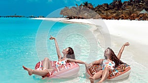 Summer Vacation. Enjoying suntan two women resting on donut float mattress in turquoise water on exotic beach. Maldives island