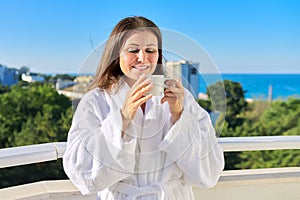 Happy middle-aged female in white bathrobe on balcony of hotel in seaside resort town