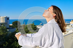 Happy mature female in white bathrobe on balcony of hotel in seaside resort town