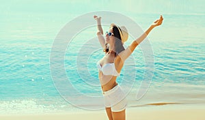 Summer vacation, happy smiling woman in bikini swimsuit and straw hat raising her hands up on the beach on sea coast background on