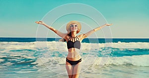 Summer vacation, happy smiling woman in bikini swimsuit and straw hat raising her hands up on the beach on sea coast background