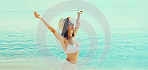 Summer vacation, happy smiling woman in bikini swimsuit and straw hat raising her hands up on the beach on sea coast background on