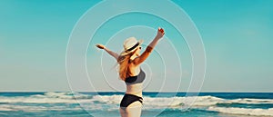 Summer vacation, rear view of beautiful happy slim woman in bikini swimsuit and straw hat raising her hands up on the beach on