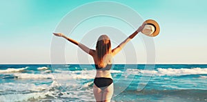 Summer vacation, rear view of beautiful happy slim woman in bikini swimsuit and straw hat raising her hands up on the beach on