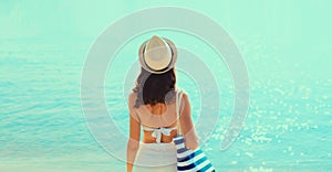 Summer vacation, rear view of beautiful young woman with bag in bikini swimsuit and straw hat on the beach on sea coast background