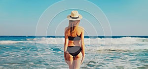 Summer vacation, rear view of beautiful young woman in bikini swimsuit and straw hat on the beach on sea coast