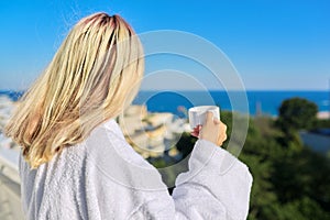 Young woman in bathrobe enjoying cup of coffee and sunny landscape of sea resort, back view