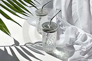 Summer vacation still life scene. Cocktail glass cups with silk fabric and palm shadow on white background.