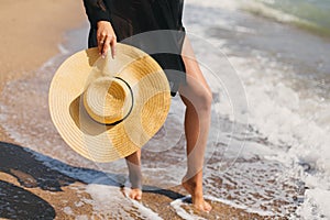 Summer vacation. Stylish hat in hands of carefree woman walking in sea waves on sunny beach, close up. Young fashionable female