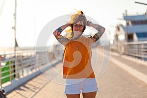 Summer vacation. Young happy Caucasian woman with tattoo smiling and posing on pier. Concept of freedom and happiness