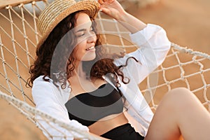 Summer vacations concept, Happy woman with black bikini, hat and white shirt relaxing in hammock on tropical beach at sunset