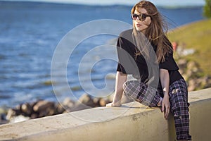 Summer Vacations.  Relaxing Caucasian Blond Female Girl in Street Clothing Posing Outdoors While Sitted On Shoreline Outdoors At