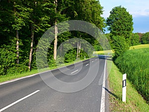 Summer view on empty automobile road with kilometer post among green fiels and forest trees. Empty car road. European car travel c