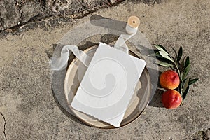 Summer wedding stationery. Blank greeting card mockup on ceramic plate in sunlight. Silk ribbon, peach fruit and olive