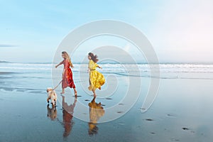 Summer. Women On Dog-Friendly Beach. Stylish Models In Boho Dresses Running Barefoot With Dog Along Coastline