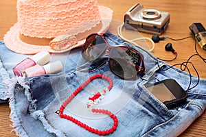 Summer women's accessories: red sunglasses, beads, denim shorts, mobile phone, headphones, a sun hat, camera, nail polish