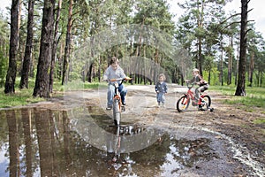 In the summer in the woods playing small children, they go through a puddle on a bicycle.