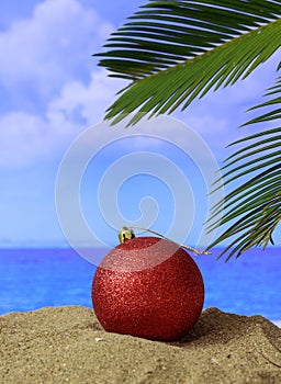 Summer xmas holidays concept. Christmas ball on sandy beach with palm tree, blue sea and sky background