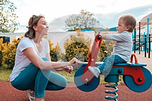 Summertime and family holidays. A young smiling Caucasian mother plays with a child on the playground, riding a baby on