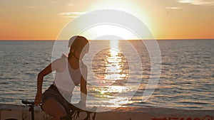 Summertime mood shot of young woman cyclist in stylish outfit is riding bicycle along beach promenade, pedalling in sunset light.
