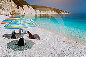 Sun beach umbrellas on a pebble beach with calm azure sea water, white cliff rocksy coastline and clear blue sky in