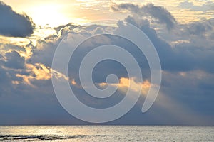 Sun with Bright, Golden Yellow and Orange Aura, behind Dark Clouds and Sunlight falling on Calm Sea Water through Clouds in Sky