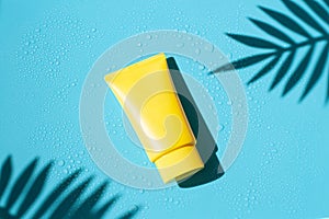 Sun cream yellow tube on the blue wet background with palm leaves shadows. Summer vacation cosmetics concept.