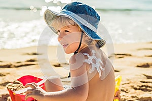 The sun drawing sunscreen ,suntan lotion on baby boy back. Caucasian child is sitting with plastic container of sunscreen and toys