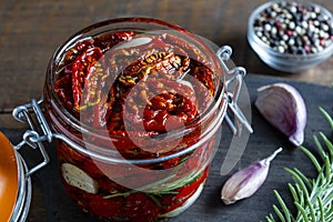 Sun-dried red tomatoes with garlic, green rosemary, olive oil and spices in a glass jar on a wooden table. Rustic style, closeup