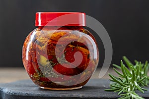 Sun-dried red tomatoes with garlic, green rosemary, olive oil and spices in a glass jar on a wooden table. Rustic style, closeup