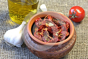 Sun-dried tomatoes with spices and garlic in a clay pot. Nearby is a bottle with olive oil, tomatoes and garlic