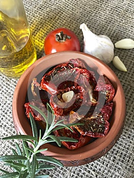 Sun-dried tomatoes with spices and garlic in a clay pot. Nearby is a bottle with olive oil, tomatoes, rosemary and garlic