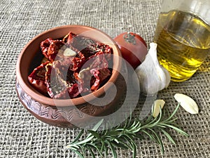 Sun-dried tomatoes with spices and garlic in a clay pot. Nearby is a bottle with olive oil, tomatoes, rosemary and garlic