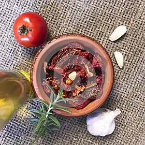 Sun-dried tomatoes with spices and garlic in a clay pot. Nearby is a bottle with olive oil, tomatoes, rosemary and garlic