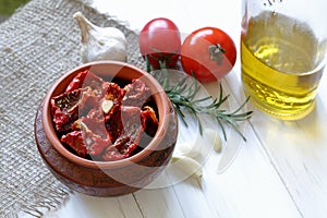 Sun-dried tomatoes with spices and garlic in a clay pot. Nearby is a bottle with olive oil, tomatoes, rosemary and garlic