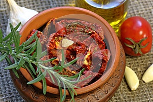 Sun-dried tomatoes with spices and garlic in a clay pot. Nearby is a bottle with olive oil, tomatoes, rosemary and garlic