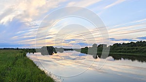 The sun has hidden behind the trees growing on the river bank. The colorful cloudy sky at sunset in summer is reflected in the wat