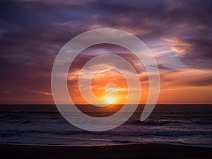 Sun on horizon at sunset with blue orange gradient sky and dark dramatic clouds over ocean