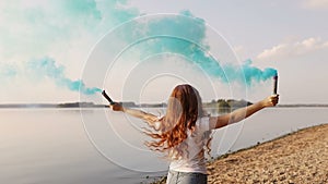 Sun-kissed freckled redhead girl with blue colorful smoke is running on bank of river