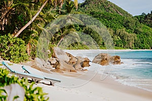 Sun lounger at secluded tropical beach. Ocean waves rolling to shore with white sand and coconut palm trees. Travel