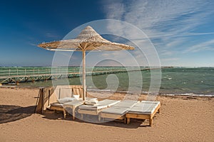 Sun lounger under the beach umbrella with view at the sea coast. Blue sky and white clouds over a resort beach.