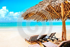 A sun lounger under an umbrella on the sandy beach by the ocean and cloudy sky. Vacation background. Idyllic beach landscape in