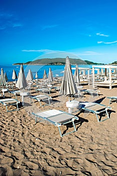 Sun loungers and umbrellas on public beach at summer sunny mornin. Skojl island background. Calm sea and blue sky Becici,